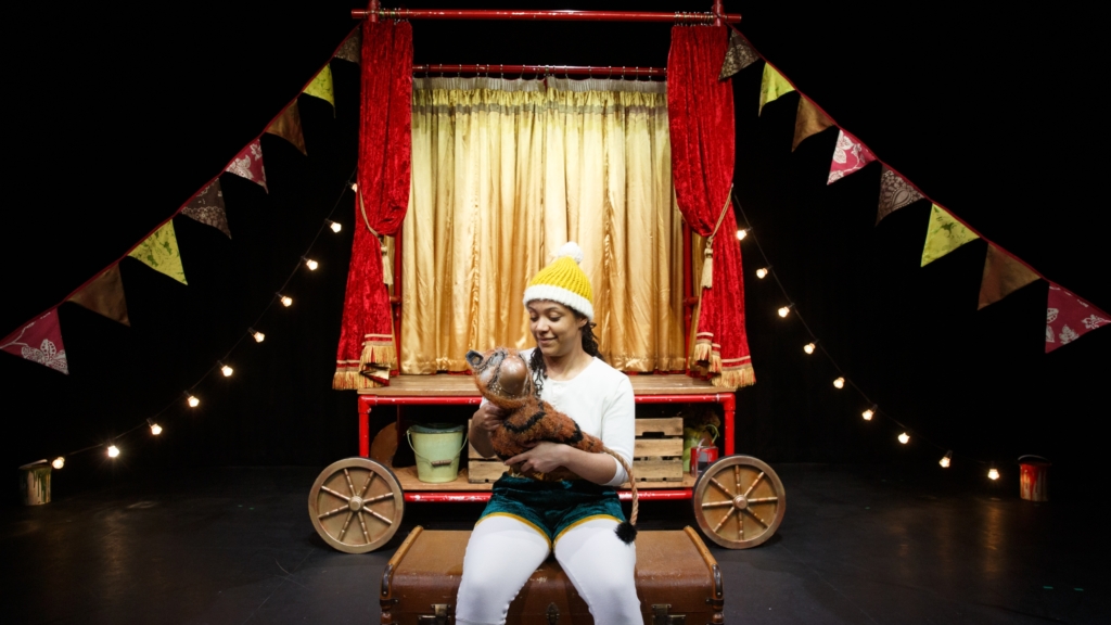 A person wearing a yellow and white knitted hat sat on a storage chest holding a prop of a baby tiger on stage.