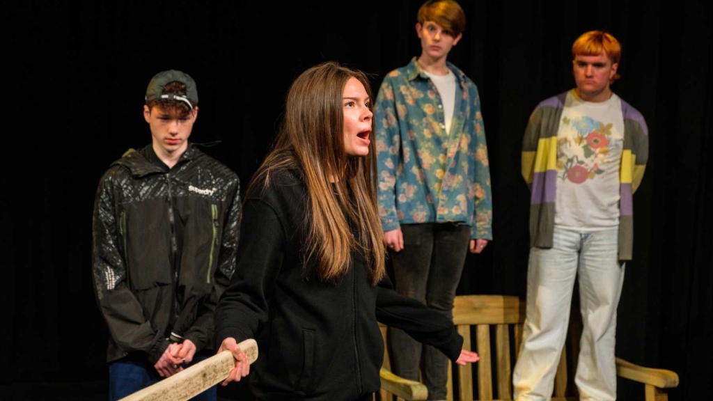 A group of 4 young performers onstage. Two performers are stood in the background on a wooden bench. A performer in the foreground holds a wooden plank in their hand, standing in front of a guilty looking performer.