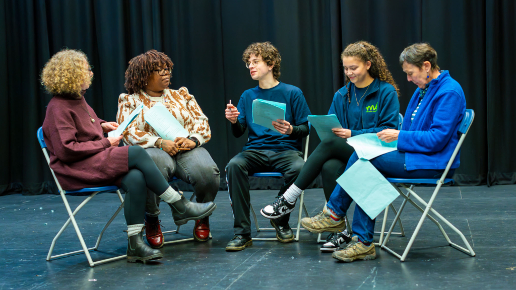 A group of people rehearsing their performance, sat on blue fold up chairs.