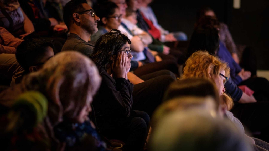 A long shot of the audience looking engaged at a performance. One audience member has her hands at the sides of her face.