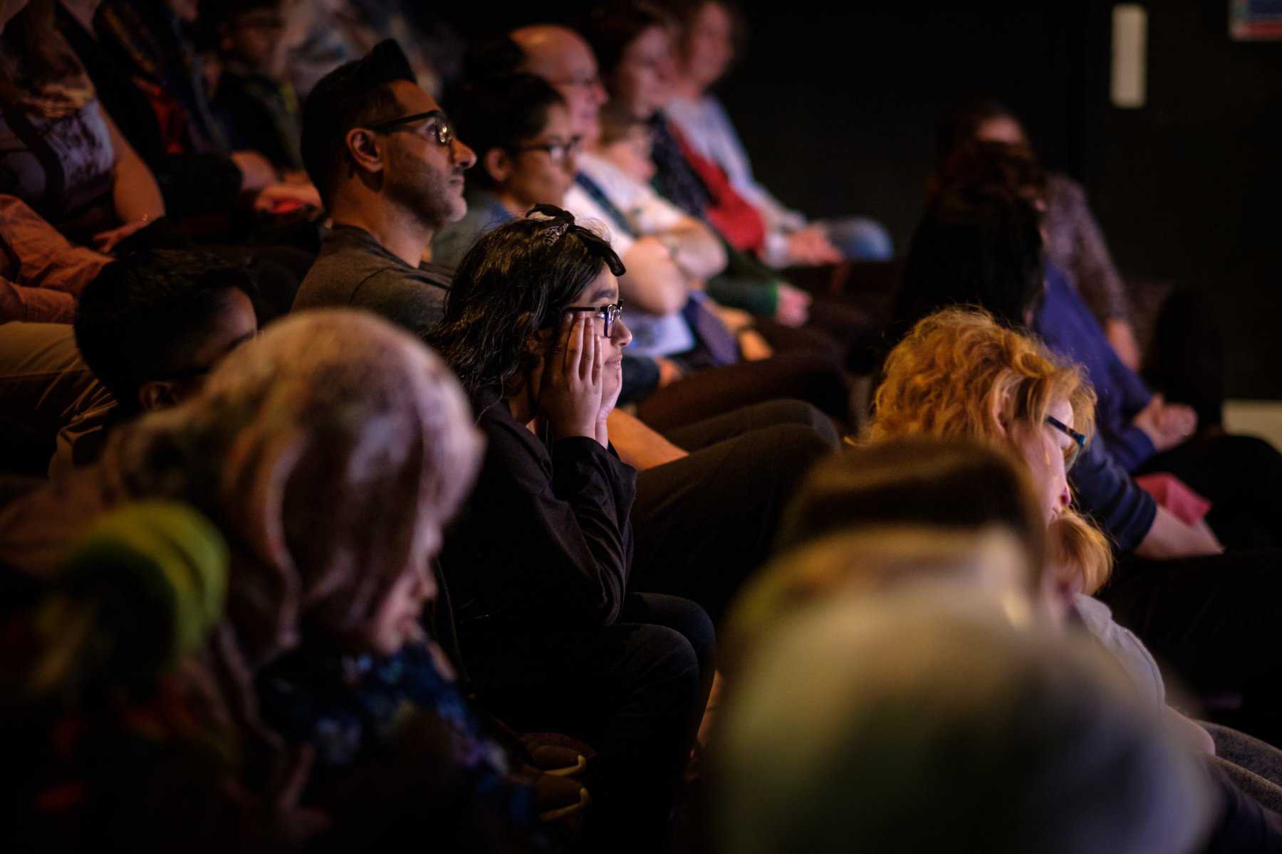 A long shot of the audience looking engaged at a performance. One audience member has her hands at the sides of her face.