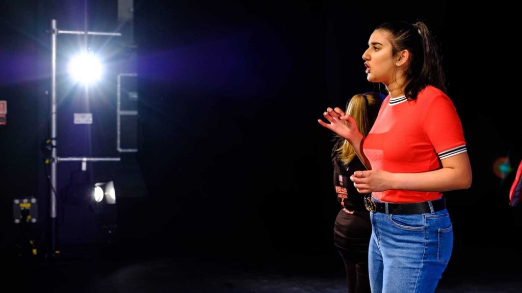 A performer on stage performing a monologue or singing. Their lips are parted and they look onward at the crowd.