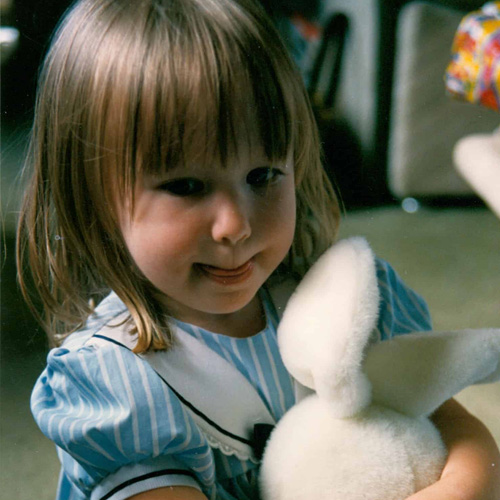 A medium close up photograph of Laura Rodwell as a child, holding a white stuffed bunny.