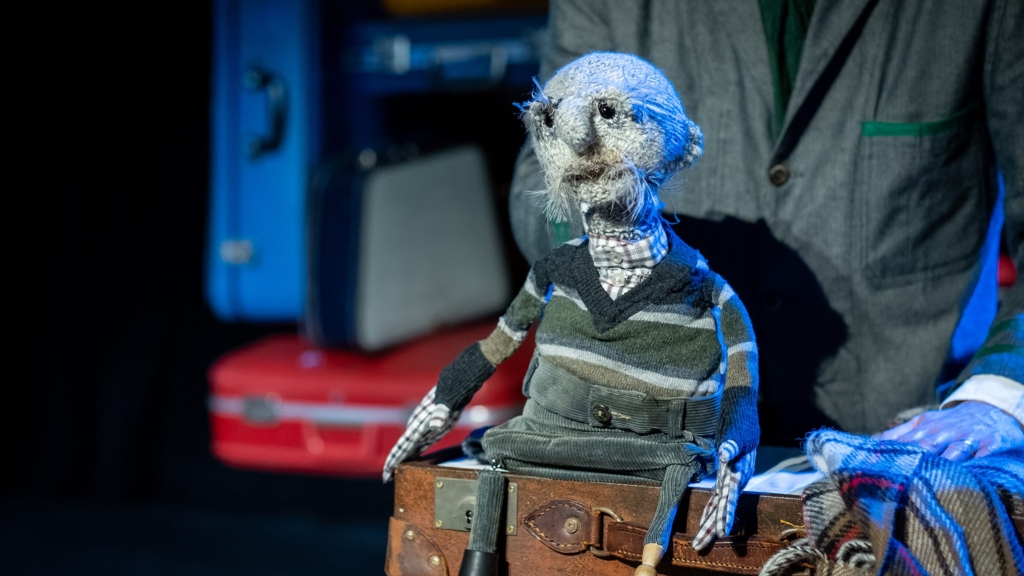 A knitted grey puppet man on stage wearing a stripey jumper, sat on a brown leather suitcase.
