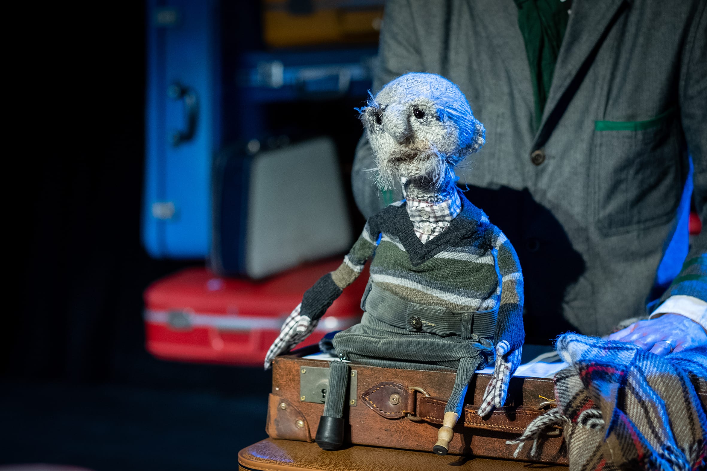 A knitted grey puppet man on stage wearing a stripey jumper, sat on a brown leather suitcase.