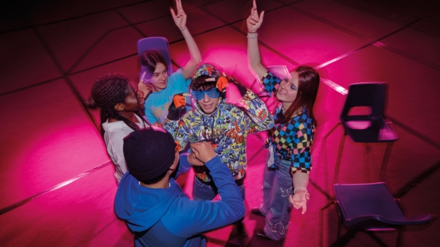 A group of young people stood up dancing in a circle. There are blue plastic chairs surrounding them.