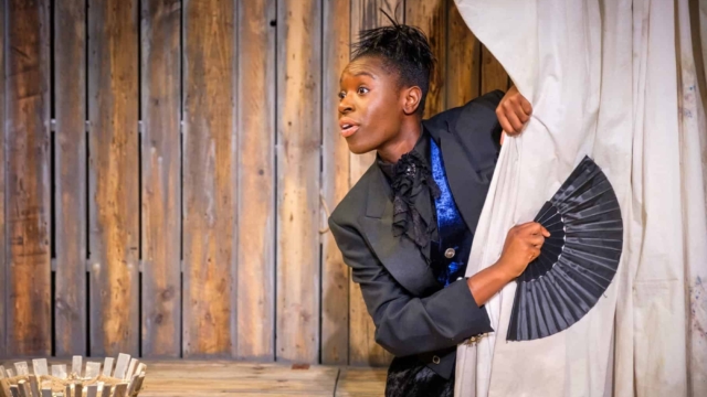 A performer on stage wearing a black jacket in front of a wooden hut, peering around a beige curtain whilst holding a black hand fan.