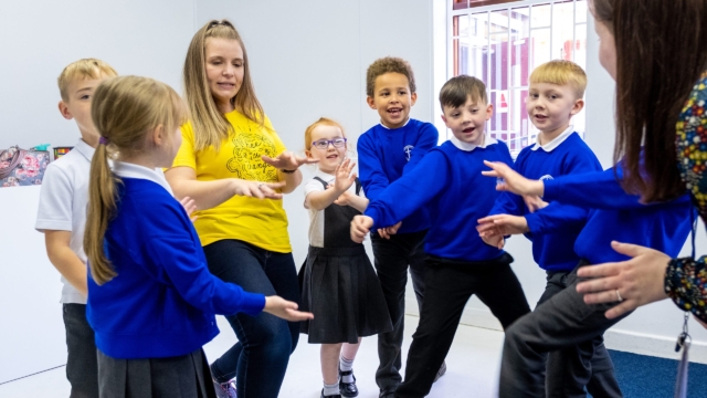 A group of primary school pupils in blue jumpers stood with an adult wearing a yellow t-shirt. They are stood in a circle with their hands inward.