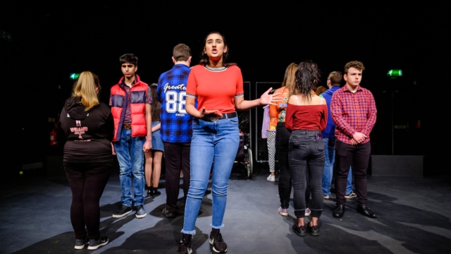 A group of young performers on a stage in different formations. Some people face onwards, whereas others have their backs facing the camera.