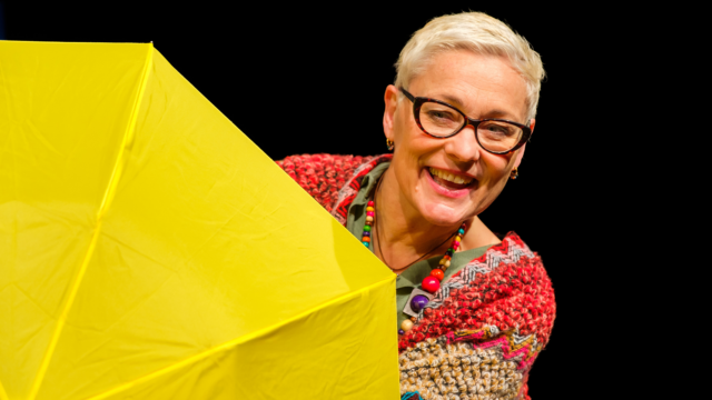 A performer on stage smiling behind a yellow umbrella.