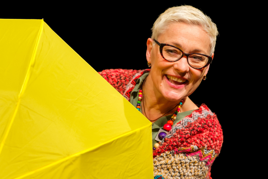 A performer on stage smiling behind a yellow umbrella.