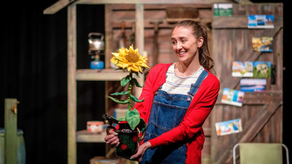 A performer onstage stood outside a wooden shed prop, holding a felt sunflower prop.