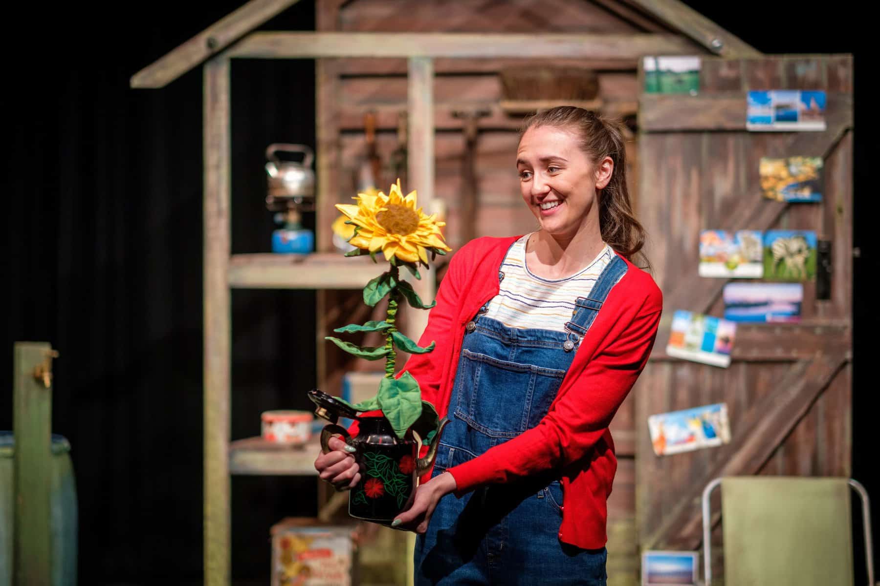 A performer onstage stood outside a wooden shed prop, holding a felt sunflower prop.