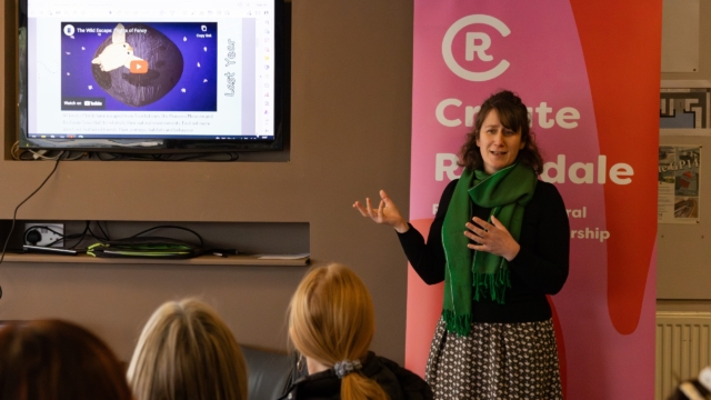 A speaker stood up giving a talk in a classroom environment with listeners who are sat down. There is a TV showcasing the YouTube video