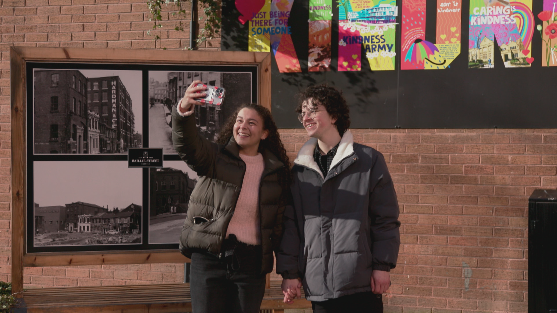 A young couple holding hands outside, stood in front of a brick wall and taking a picture together on a mobile phone.