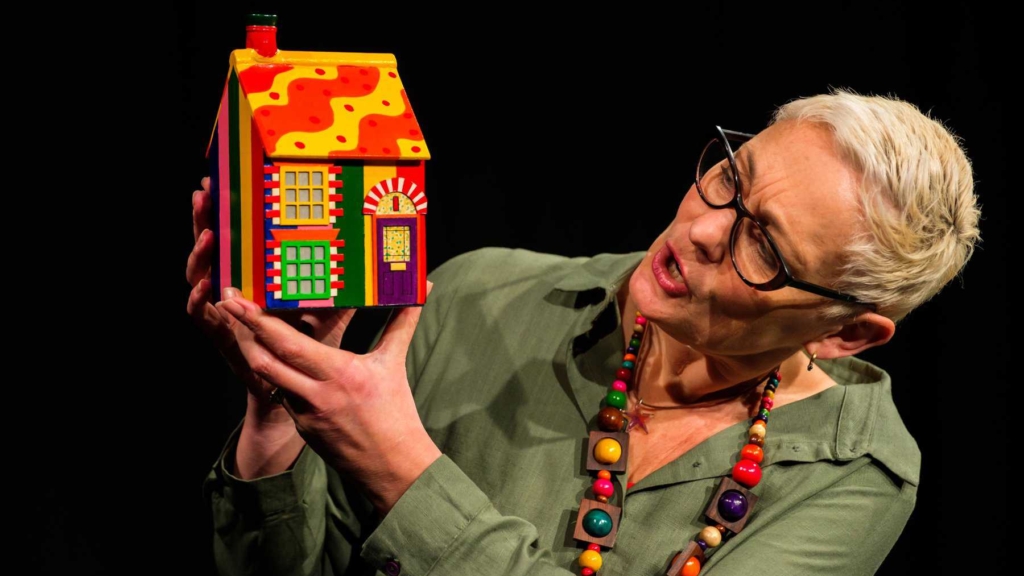 A speaker holding a minature model house prop in their hands. It is painted in many bright colours.