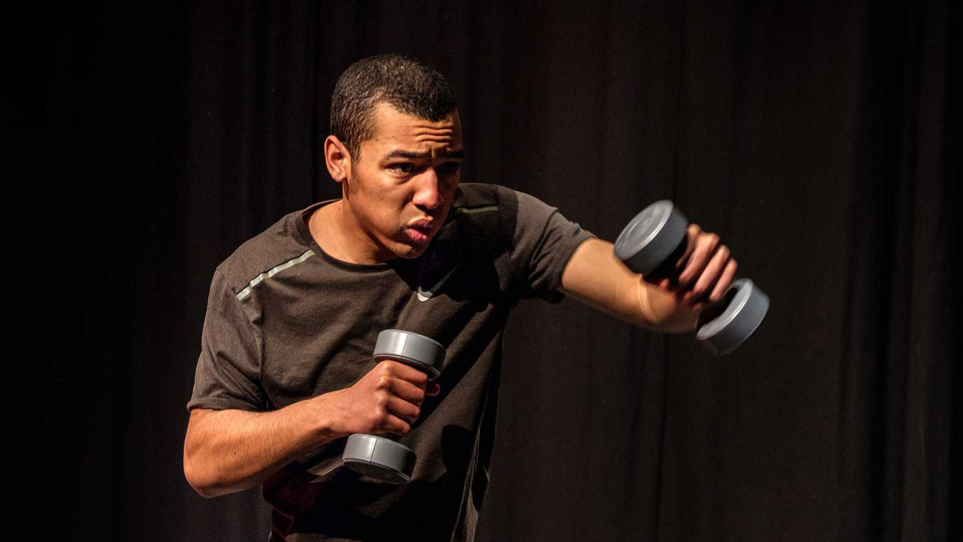 An adult male performer on stage wearing a grey Nike t-shirt and exercising with small grey dumb bells in his hands.