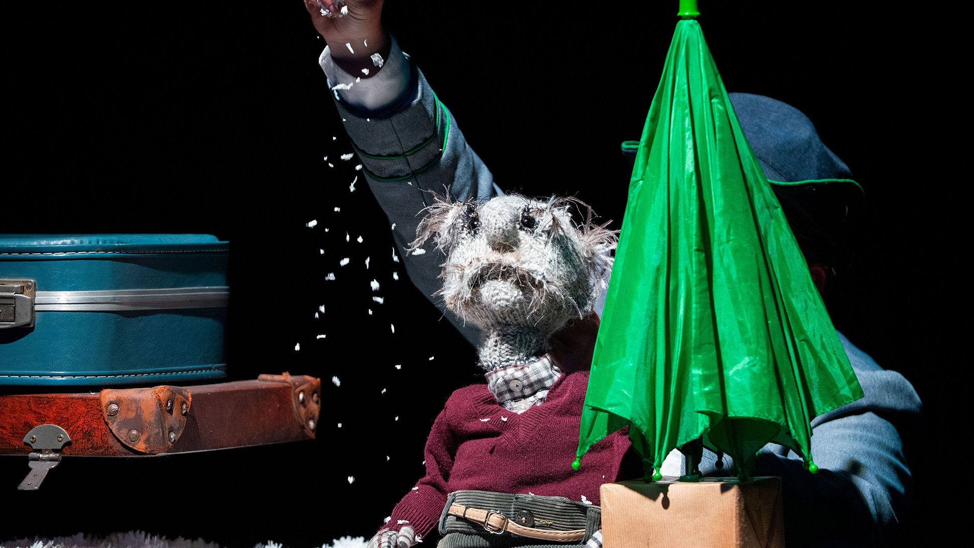 A knitted grey puppet man on stage wearing a maroon jumper, sat on a brown leather suitcase next to a folded green umbrella, as paper snow falls above him.