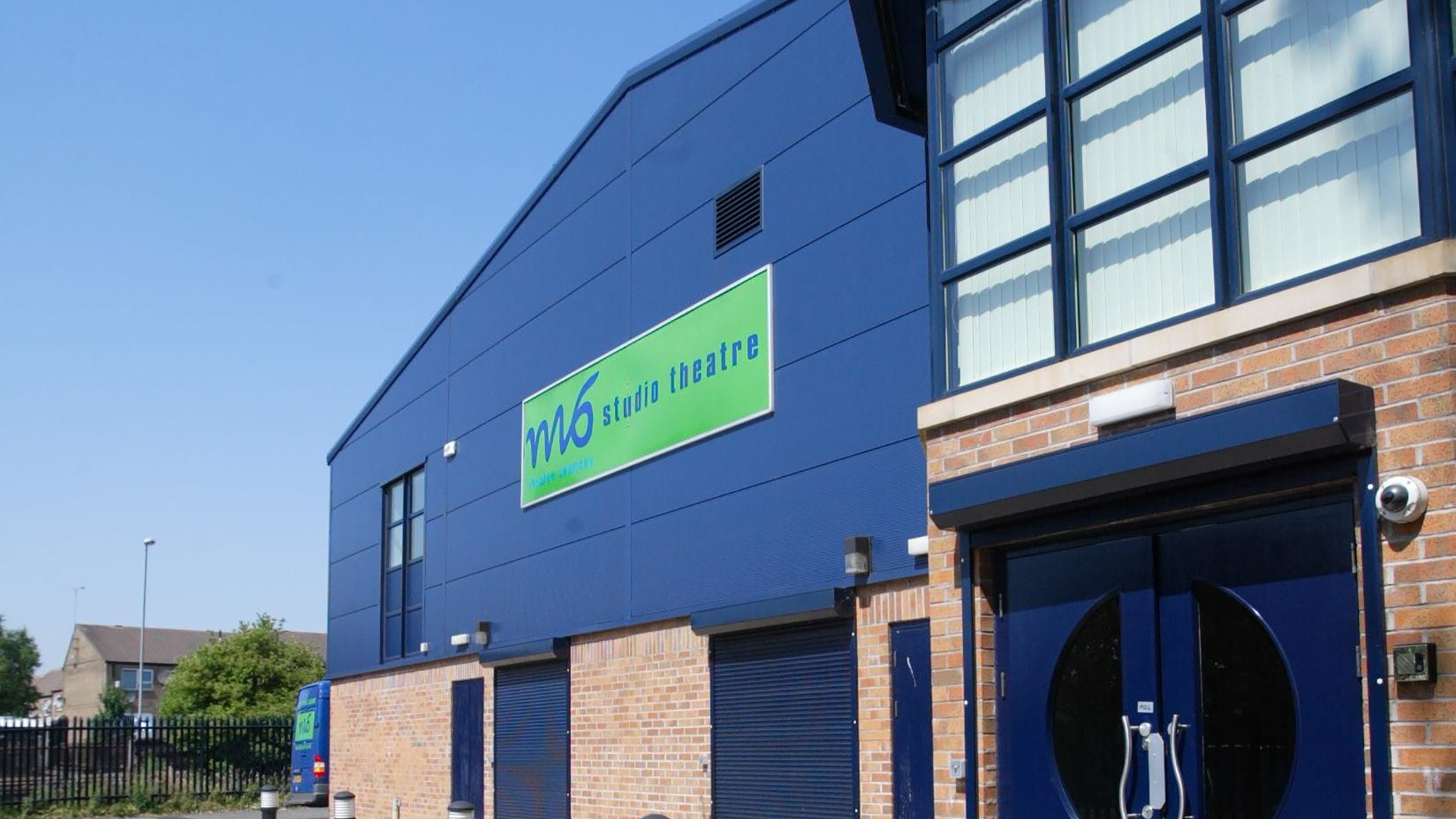 The outside of the blue M6 Studio Theatre building. Featuring red bricks and a green M6 Studio Theatre sign.