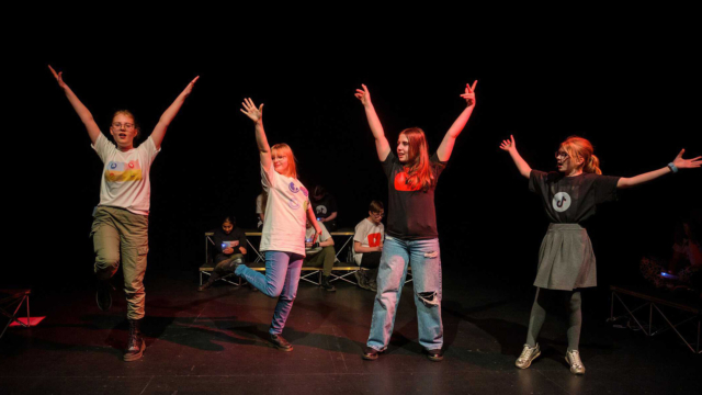 A group of performers on stage in different poses with their arms stretched upwards. The performers have t-shirts with the logos of different social media networks on them e.g. YouTube and TikTok.