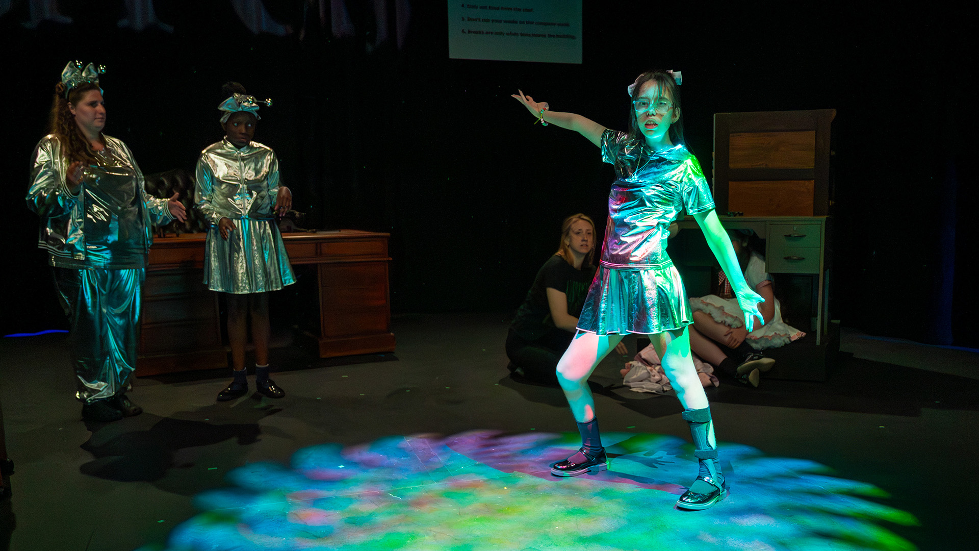 A group of young performers on stage mid performance. They are all dressed in shiny silver outfits.