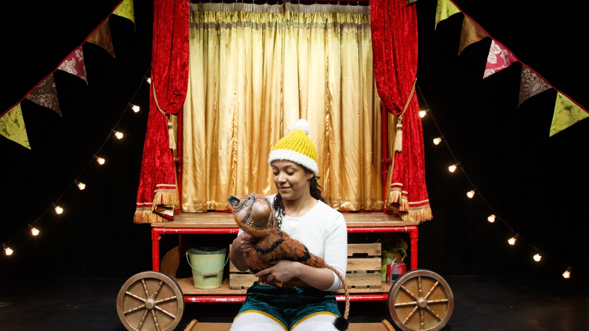 A person wearing a yellow and white knitted hat sat on a storage chest holding a prop of a baby tiger on stage.