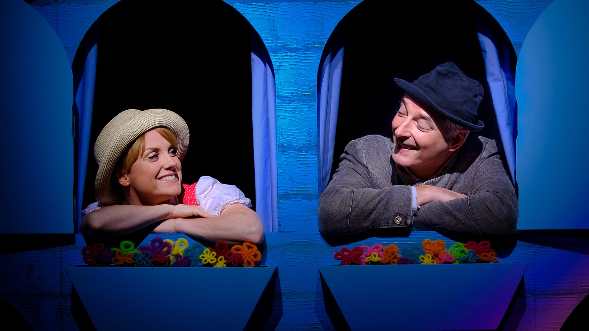 A couple of performers on stage using a prop of a cardboard house that has two windows. They stand with their arms folded over the cardboard window frames, looking at each other happily. There are felt flower beds underneath them.