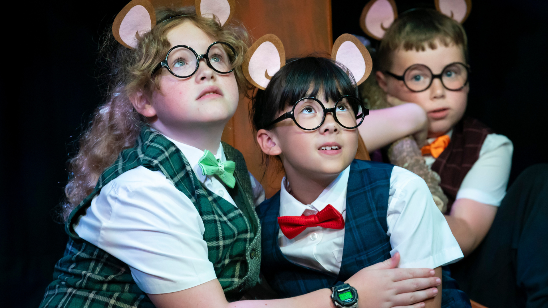 A close up of young performers on stage dressed as mice wearing black circular glasses, checkered waistcoats, bow ties and brown paper mouse ears.